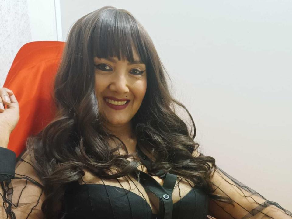 SweetFoxyne with wavy brown hair, smiling, wearing black sheer top, showing cleavage, sitting on red chair, holding her hand near her face, cute, attractive, sexy.