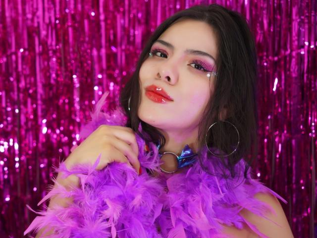 MaraCortes com uma echarpe de penas roxas, maquiagem rosa brilhante, brincos de argola e um colar azul, exibindo lábios vermelhos brilhantes, com um fundo de glitter rosa. Sua mão está perto do rosto..