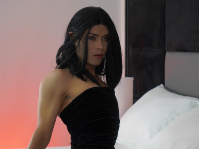 AlexaDelys with black hair, wearing a strapless black velvet dress, showing her ass, sitting on a bed with white pillows, looking serious, in a dimly lit room.