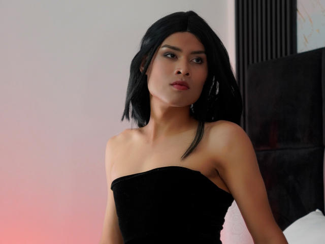 AlexaDelys has black shoulder-length hair, a serious expression, and is wearing a strapless black dress, exposing her bare shoulders and upper chest. Her ass is partially visible.