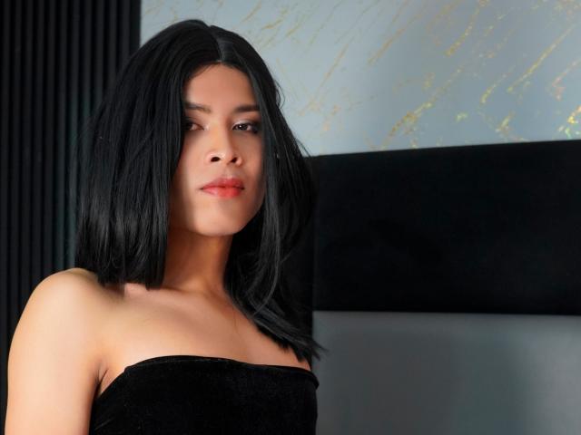 AlexaDelys with black shoulder-length hair, light skin, and red lipstick, wearing a black strapless top, looking slightly to the side with a calm expression.
