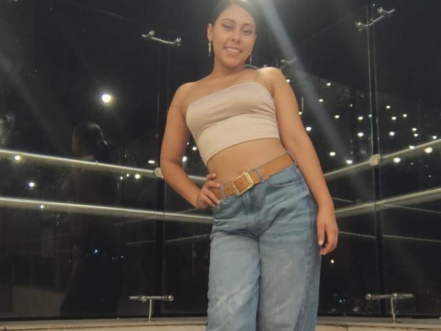 MarilynHills, standing, smiling, wearing a beige strapless top, blue jeans with a brown belt, and earring, hand on hip, light reflecting on glass behind her.