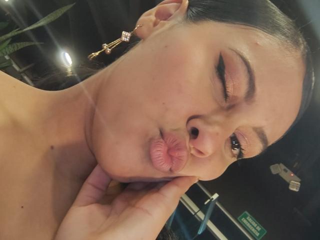 MarilynHills, with puffy pink lips, winking, holding her chin, has dark brown hair, wearing gold earrings, in a selfie. Her nose is slightly upturned.
