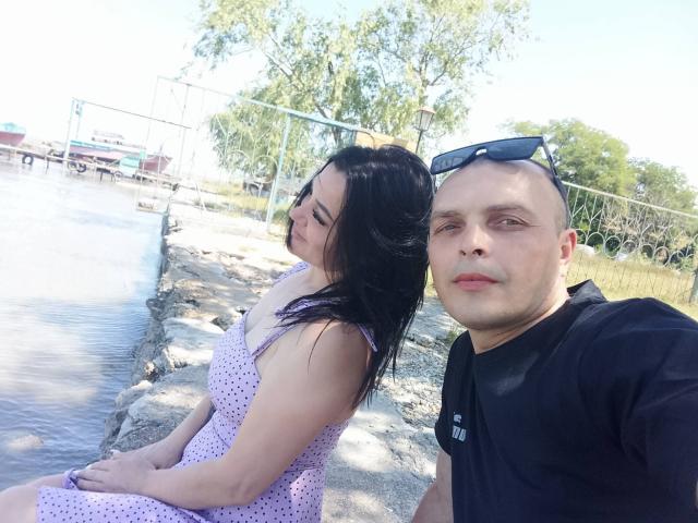 Alica e Barry, Alica con i capelli neri e un vestito rosa a pois, e Barry con la testa calva e una camicia nera, seduti vicino all'acqua, una giornata di sole, cielo blu..