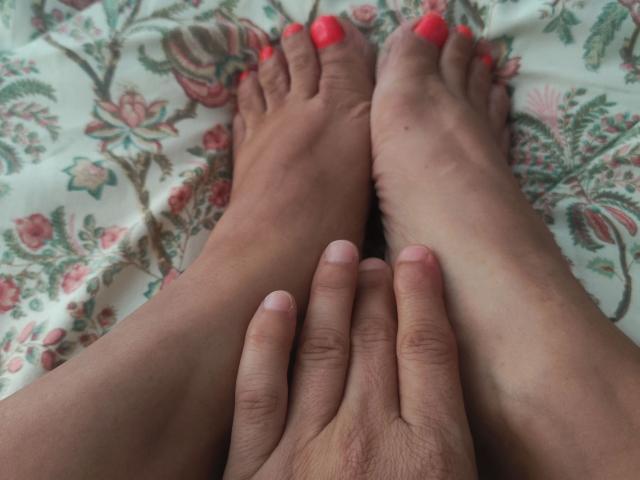 CleopatraUnica-hot's feet with neon pink toenails, hands resting on her lower legs. Her skin is light brown, nails are neatly trimmed. She's lying on floral-patterned bedsheet.