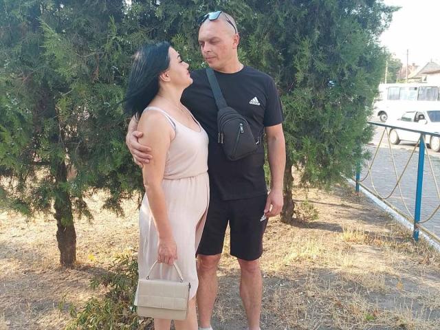 Alica e Barry, com cabelo preto, vestido rosa, segurando uma bolsa, em pé ao lado de Barry, careca, camisa preta, shorts pretos, bolsa preta, perto de árvores, durante o dia, olhando um para o outro..