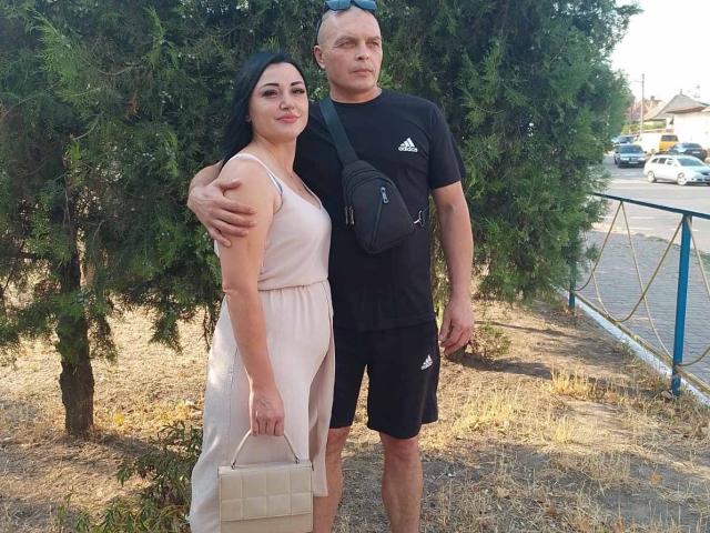 Alica et Barry, avec ses cheveux noirs et sa robe rose, se tiennent à côté de Barry, qui porte une chemise noire et un short, et a un bras autour d'elle. Elle tient un sac beige, tandis que lui a un sac noir..