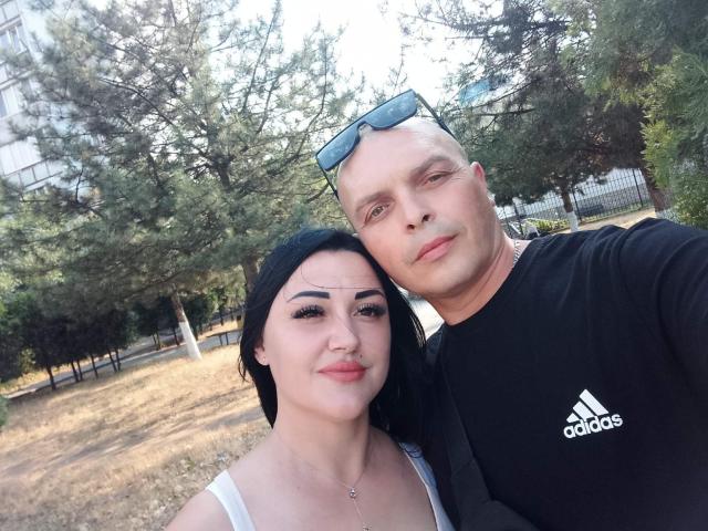 AlicaAndBarry with black hair and small tits, wearing a white top, next to a bald man with blue sunglasses on his head, in a black Adidas shirt.