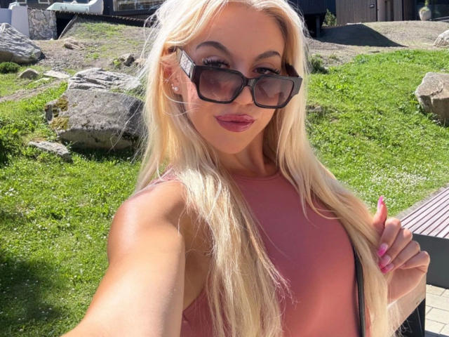 CarolyneMarian-hot with long blonde hair, pink lipstick, and black sunglasses, wearing a pink sleeveless top, holding her hair, outdoors, with a grassy background and rocks.