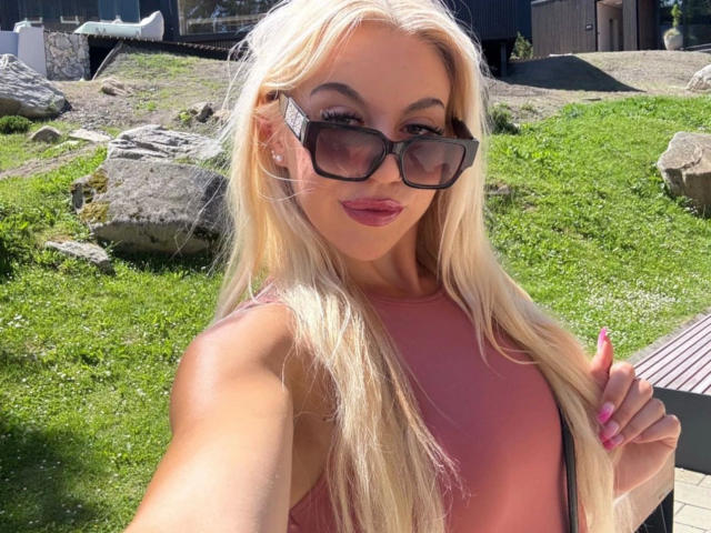 Blonde CarolyneMarian-hot with long hair, pink lips, and pink top, wearing black sunglasses, smiling, holding hair, and showing pink nails, standing in a sunny grassy area.