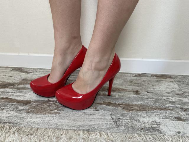 Reiza's shiny red high heels with a glossy finish, standing on a wooden floor, showing her pale legs and slender ankles.