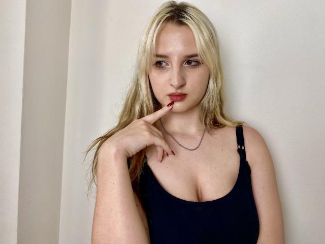 Reiza with blonde hair, pale skin, and red lipstick, wearing a black tank top, has her finger on her red-painted lip, showing off her small tits.
