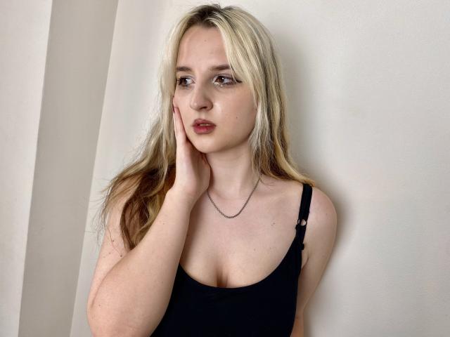 Reiza with blonde and brown hair, black tank top, and hand on cheek, showing her tits, pale skin, and pink lips, wearing a thin necklace, in a photo.