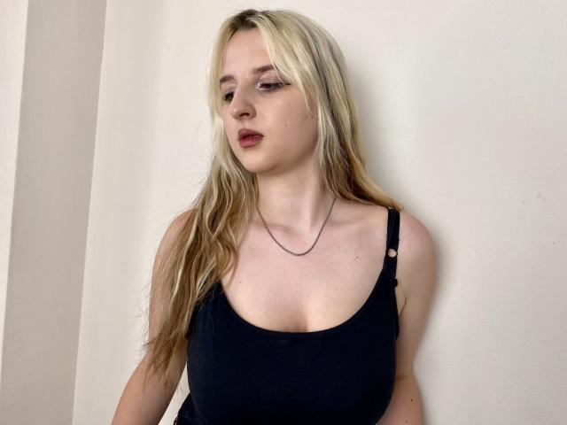 Reiza has blonde hair, wearing a black tank top, showing her tits, with a subtle necklace, and a pouty expression, against a plain white wall.