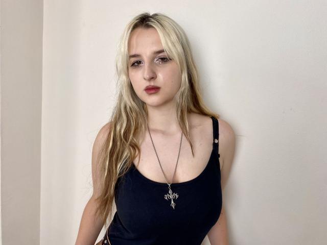 Reiza with blonde hair, wearing a black tank top, showing her boobs, has a necklace with a small pendant, standing against a white wall, looking slightly bored.