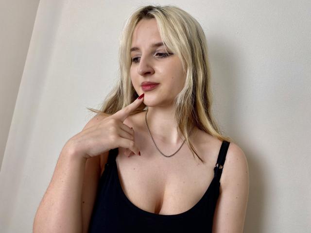 Reiza with blonde hair, black tank top, and red nails, gently touching her chin with a slight smile, showing her tits. She wears a thin necklace.