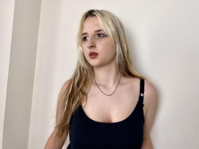 Reiza with blonde hair, black tank top, visible cleavage, and subtle makeup, looking to the side with a slight pout. Simple necklace, pale skin, and relaxed expression.