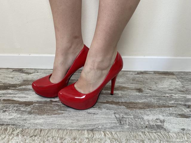 Les élégantes chaussures rouges de Reiza, avec leur finition brillante, se trouvent sur un plancher en bois recouvert d'un tapis texturé, mettant en valeur ses jambes pâles et la légère forme de sa cheville..