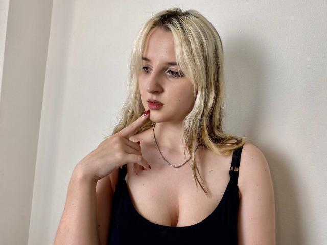 Reiza with blonde hair, black tank top, and red nails, touching her chin, looking to the side, showing her small tits and fair skin, against a white wall.