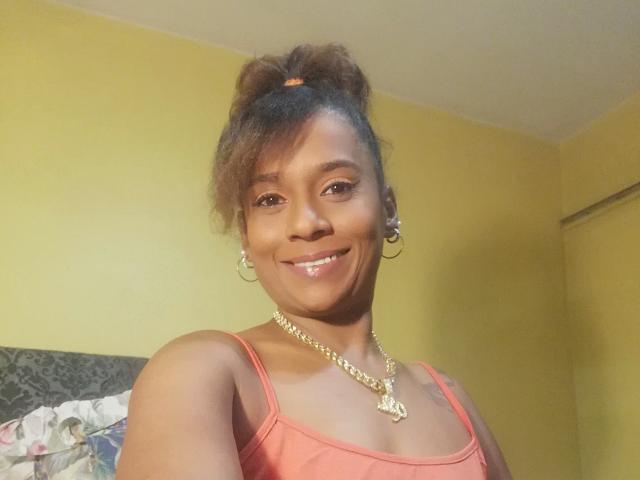 MiZzCreator, smiling with glossy lips, brown curly hair in a bun, wearing a pink tank top, gold necklace, hoop earrings, yellow wall, and a grey-patterned bed in the background.
