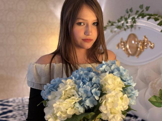 Bubochka with long brown hair, off-shoulder white and black top, holding a bouquet of blue and white hydrangeas, looking slightly to the side with a subtle smile.