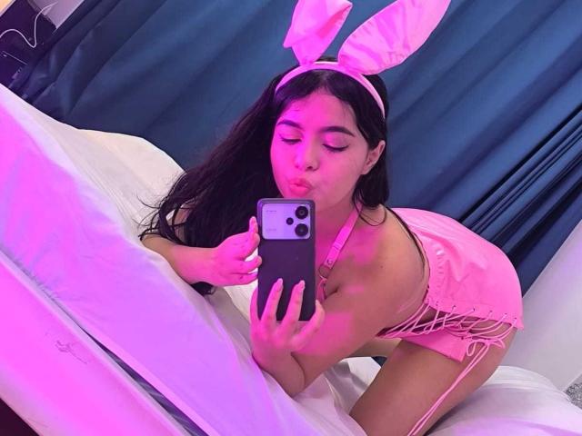 LissyCarter with pink bunny ears, kissing camera, pink lingerie, lace-up, garter, kneeling on bed, taking selfie with iPhone, pink and purple lighting, long dark hair, white bed.