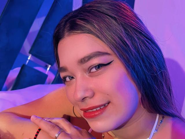 SeleneI a de longs cheveux bruns, du rouge à lèvres, et un trait d'eye-liner ailé. Elle sourit, avec des ongles peints en rouge et une bague, éclairée par des lumières violettes et bleues..