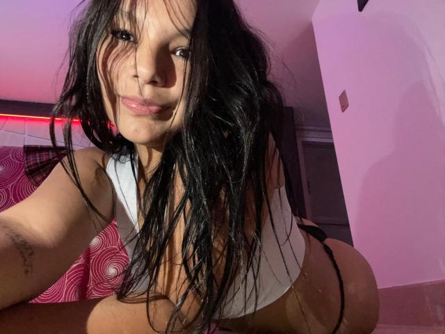 ArianaLee, avec ses cheveux noirs mouillés, son débardeur blanc et sa culotte noire, est accroupie, montrant ses fesses volumineuses et un petit tatouage sur son bras droit. L'éclairage est d'un rose doux..