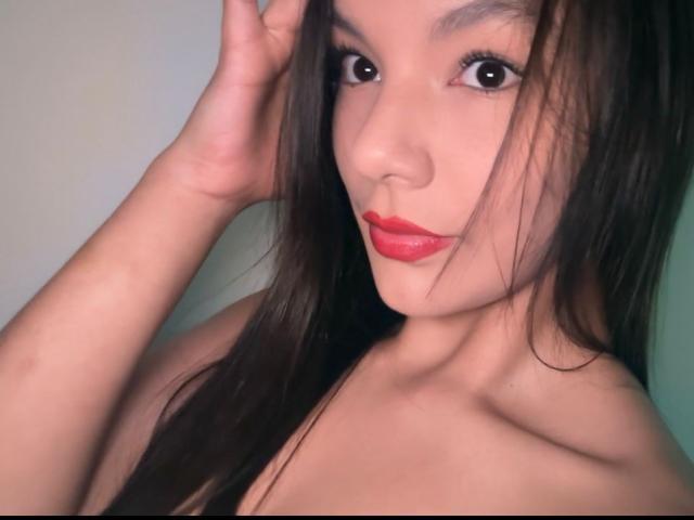 ArianaLee, com cabelos longos e pretos, batom vermelho e olhos castanhos, tocando a cabeça com uma mão, mostrando seus ombros nus. Ela tem um rosto bonito e sensual..