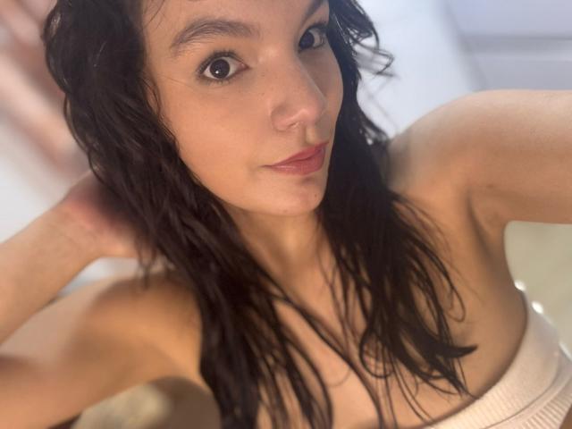 ArianaLee, avec ses cheveux noirs mouillés, son petit nez et ses yeux marrons, regarde l'appareil photo, montrant ses épaules nues et son haut blanc, qui couvre ses seins..