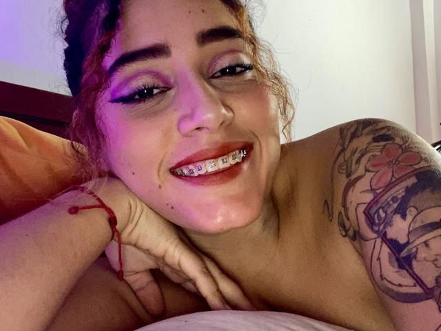 MerydaBraveI has curly brown hair, purple eyeshadow, and is smiling with metal braces showing. She's topless, revealing a tattoo on her right shoulder, and wearing a red bracelet.