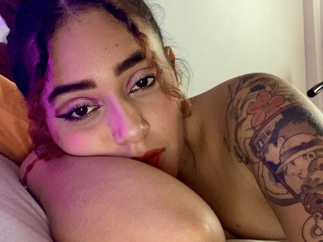 MerydaBraveI with braided hair, purple eye makeup, and tattoos, resting her head on a leg, showing her red lips and looking at the camera. Her tits are partially visible.