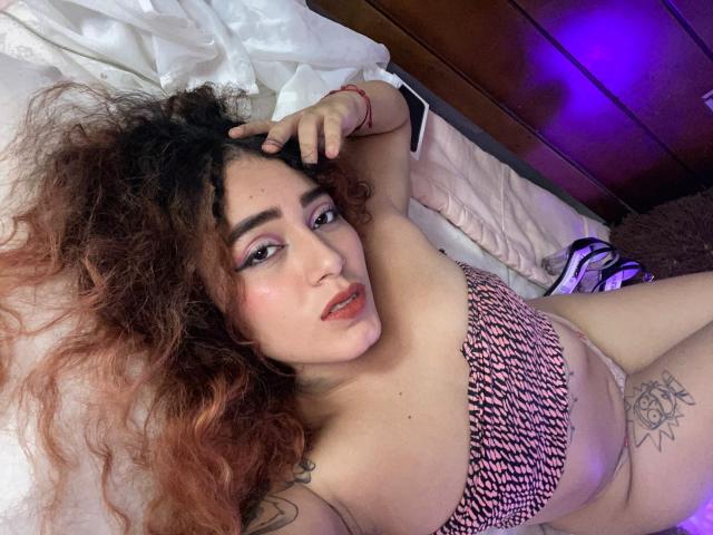 MerydaBraveI lies on a bed, wearing a pink and black striped bra, with curly brown hair, tattoos, and a seductive expression, showing her tits and a red bracelet.