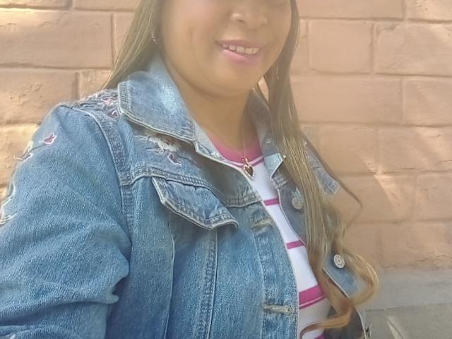 Rahel69 with long brown hair, wearing a blue denim jacket over a white and pink striped shirt, smiling with pink lips. Her tits are partially visible under the jacket.