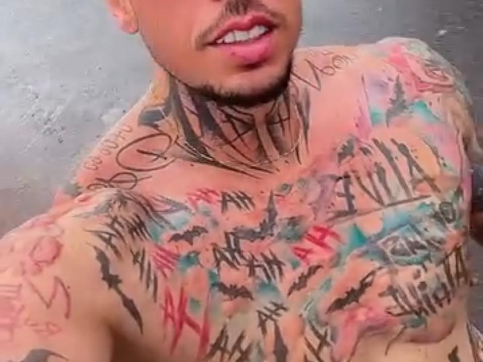 MarioStar's tattooed, shirtless upper body, with colorful ink and black script covering his chest and neck, showing a mix of words and symbols. His mouth is slightly open.