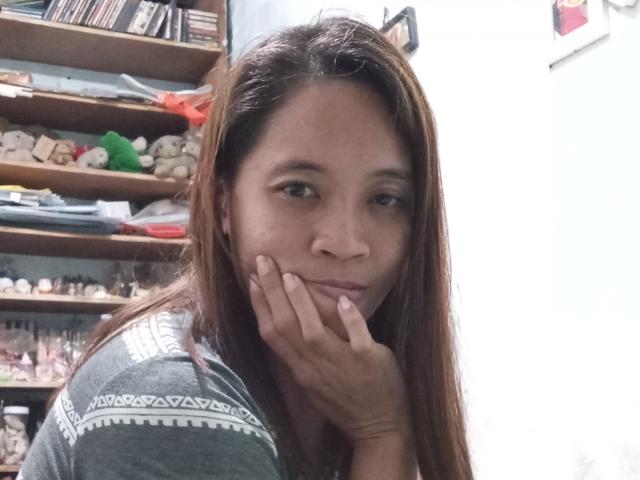 Kath69 with long brown hair, gray patterned shirt, hand near face, slight smile, looking at camera, in a cluttered room, shelves with toys and books in the background.