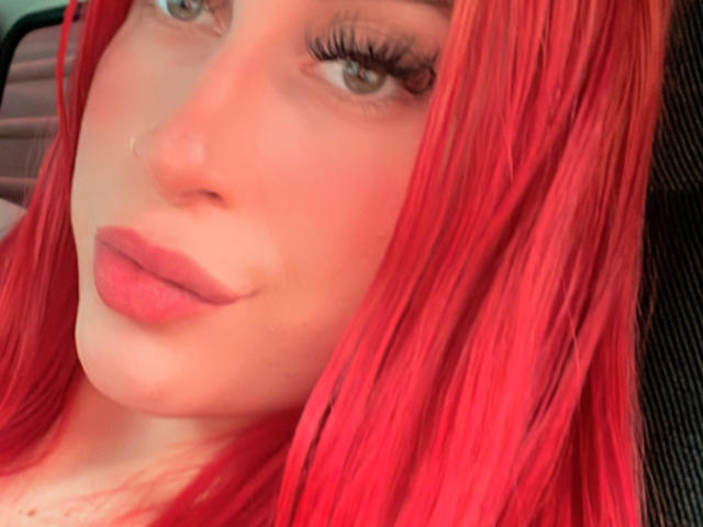 SammyViolet has red hair, green eyes, and pink lipstick. Her eyelashes are long and she's looking to the side with a slight smile on her pouty lips.