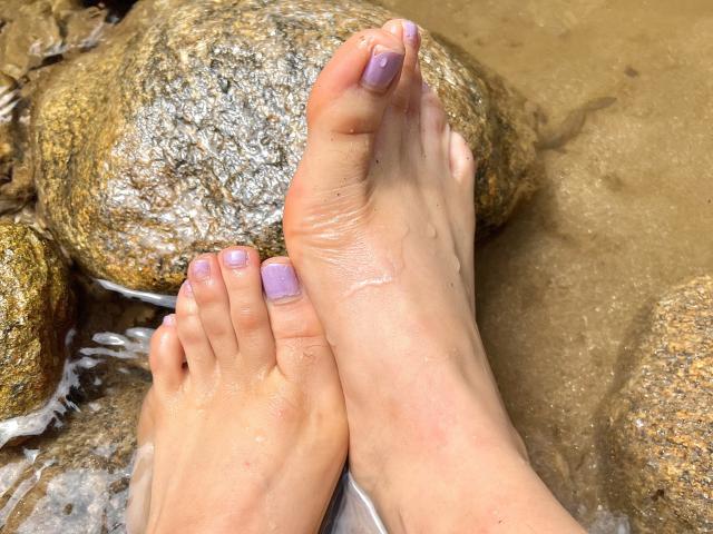 GilianRowe's feet with light purple nail polish, wet and resting on a rocky surface, showing slight sand and water. Her toes are slightly parted, showing her toes' texture.