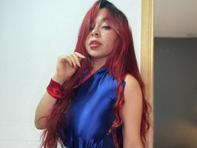 GilianRowe with long red hair, wearing a shiny blue dress, red bracelet, and pink lipstick, touching her chin, looking confident. Her tits are slightly visible under the dress.