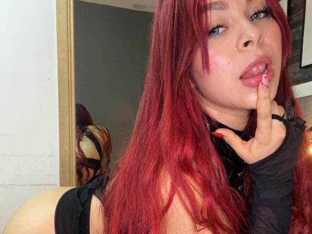 GilianRowe with red hair, biting her finger, looking at the camera. She has black gloves, shiny pink lips, and a sexy expression. Her reflection is in the mirror.