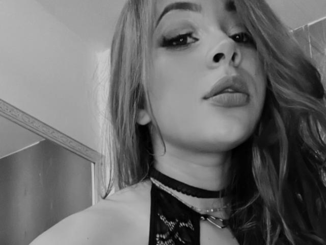 GilianRowe has long, wavy hair, thick eyebrows, and pouty lips with a black lace choker and eye makeup, looking down slightly with a sultry expression.