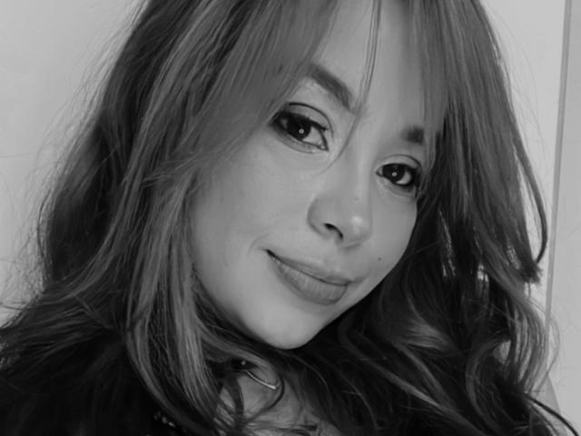 GilianRowe's face with long, wavy hair, small nose, and subtle smile in a black-and-white selfie. She has dark eyeliner, light skin, and a slight lip plump.