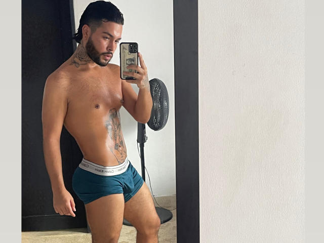 LeanPrensoom, shirtless, with tattoos, green Calvin Klein underwear, holding phone, mirror selfie, dark hair, beard, standing, leg slightly forward, black fan in background, medium build, brown skin.