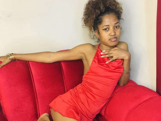 ClaraMagik in a red dress, sitting on a red velvet couch, arm stretched, hand on chest, curly hair, brown skin, small tits, subtle makeup, gold bracelet, confident expression.