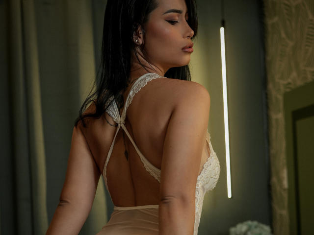 Ivannaah's back is to the camera, wearing a sheer, lace-up, backless dress, with her dark hair down, showing off her smooth skin and small tattoo on her left arm.