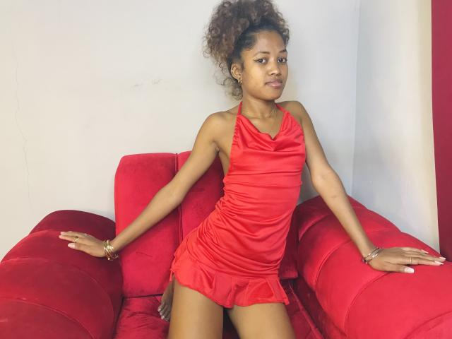 ClaraMagik with curly brown hair, wearing a red halter dress and gold bracelets, sitting on a red couch, hands on arms, looking to the side with a neutral expression.