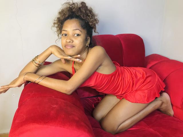 ClaraMagik, with curly brown hair, wears a red dress, kneeling on a red couch, smiling, looking at camera, with gold bracelets, and one hand touching her chin.