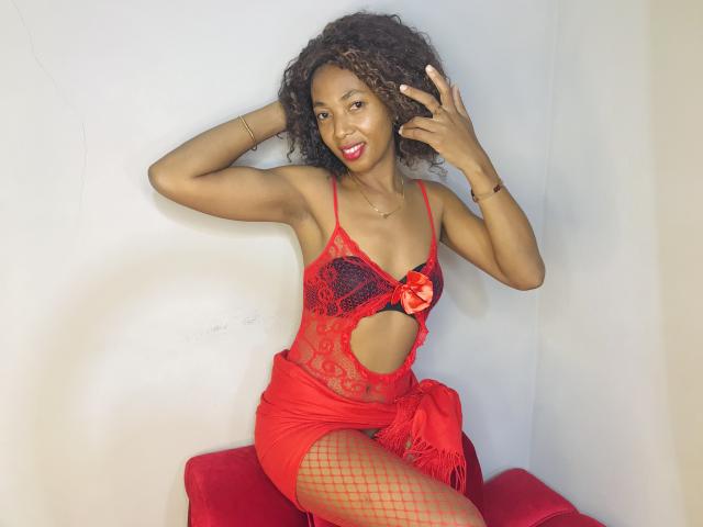 NelvaMagik with curly hair, red lace lingerie, cutout front, red fishnet stockings, red frilled skirt, sitting on red bench, smiling, hands in curly hair, red lipstick.