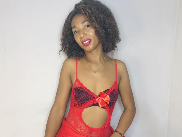 NelvaMagik with curly brown hair, red lace lingerie, and a red bow, smiling at the camera, showing her small tits and cutout in the red lingerie.