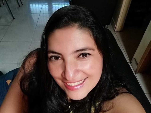 Lina69s has dark brown eyes, long black hair, and a cute smile with shiny pink lips. She's sitting on a black surface, wearing a sleeveless top, with a tile floor.